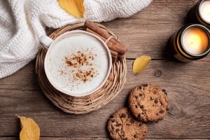 Autumn cozy composition. Cup of cappuccino coffee, chocolate chip cookies, burning candles and yellow autumn leaves on wooden background, top view. Hygge comfortable concept flat lay.